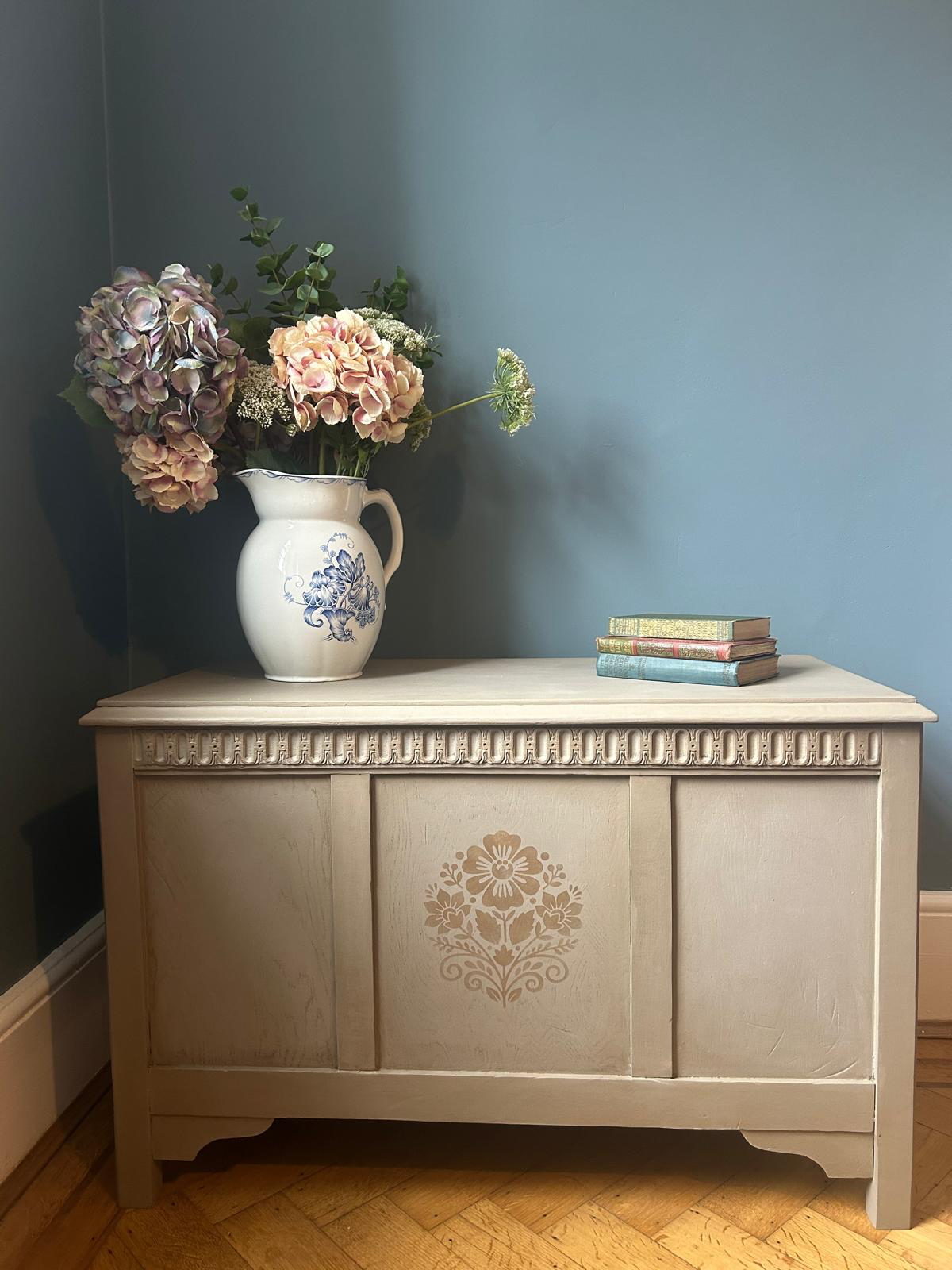 A restored pine blanket chest with a stenciled floral motif, topped with a jug of dried hydrangeas and stacked books